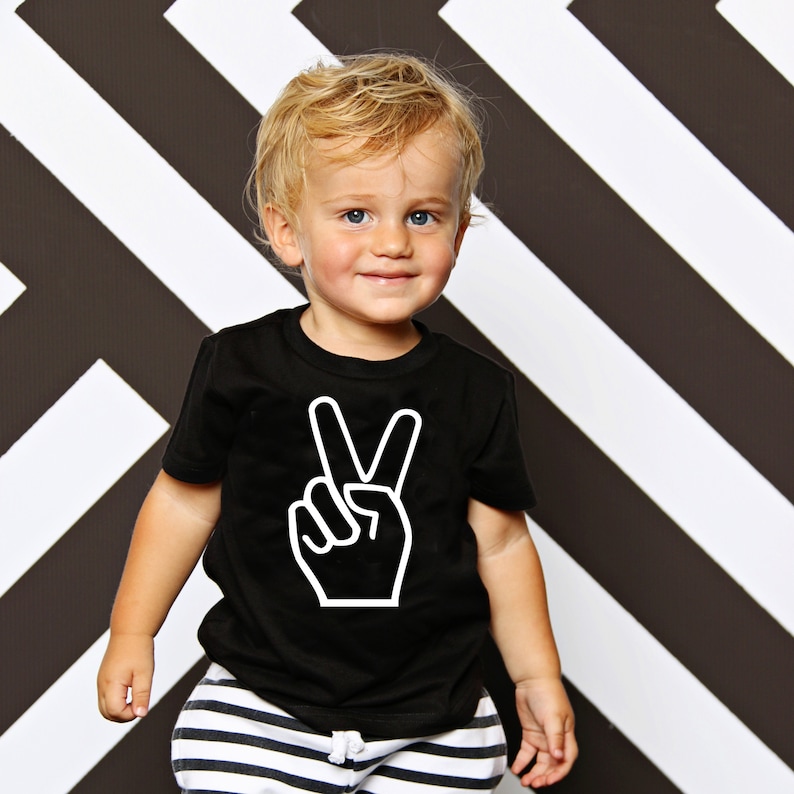 May include: A young child wearing a black t-shirt with a white peace sign graphic. The child is standing in front of a black and white striped background.