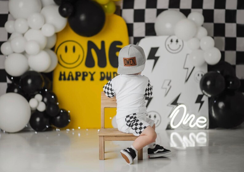 May include: A young child wearing a grey baseball cap with the name "Owen" on it sits on a wooden chair in front of a yellow and white backdrop. The backdrop features the word "One" in a black and white design and the words "Happy Dude". The child is wearing a white shirt with black and white checked sleeves and black and white checked shorts.