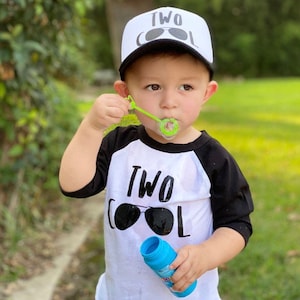 May include: A toddler wearing a white and black baseball cap with the text "TWO COOL" and sunglasses printed on it. The toddler is wearing a white and black baseball shirt with the text "TWO COOL" and sunglasses printed on it. The toddler is blowing bubbles.