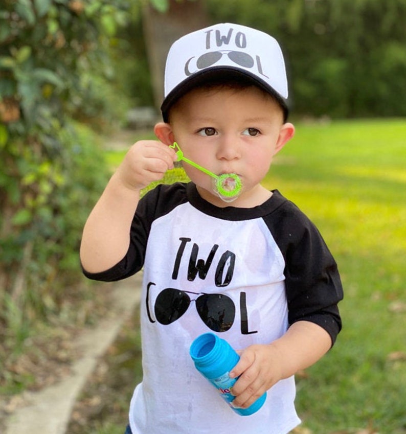 May include: A toddler wearing a white and black baseball cap with the text "TWO COOL" and sunglasses printed on it. The toddler is wearing a white and black baseball shirt with the text "TWO COOL" and sunglasses printed on it. The toddler is blowing bubbles.
