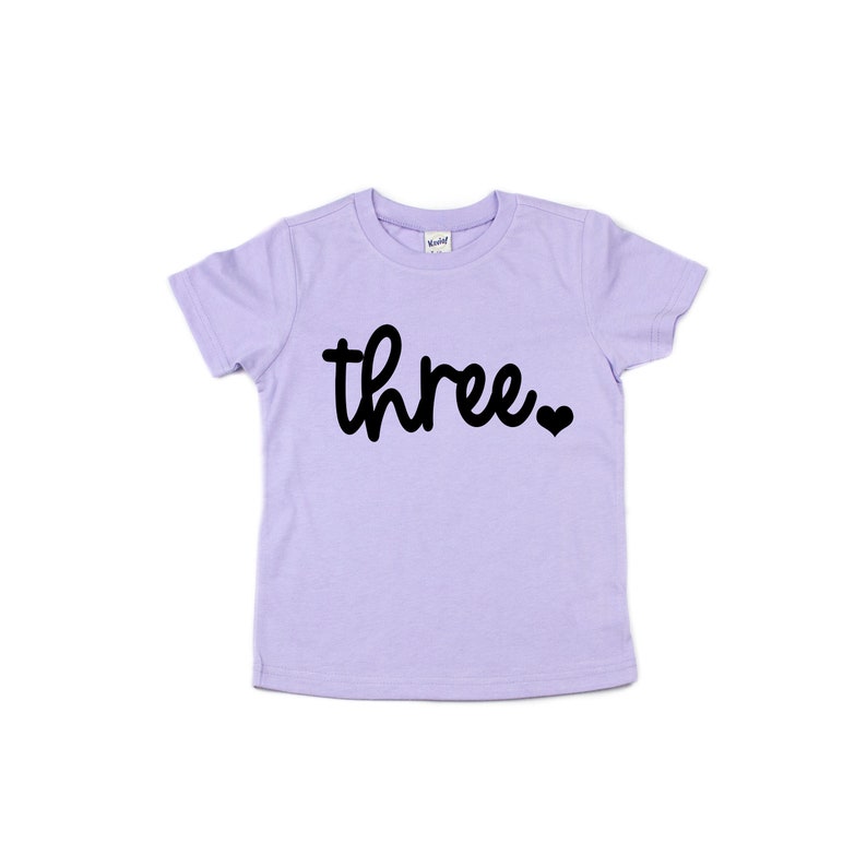 Puede incluir: Camiseta lila claro para ni&ntilde;os peque&ntilde;os con la palabra "three" en escritura negra, seguida de un peque&ntilde;o coraz&oacute;n negro. La camiseta est&aacute; hecha de material suave y est&aacute; dise&ntilde;ada para el tercer cumplea&ntilde;os de un ni&ntilde;o.
