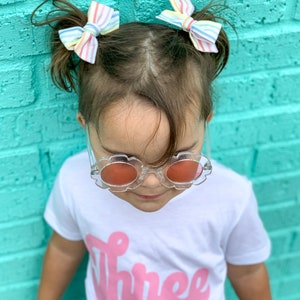 May include: A young child wearing a white t-shirt with the word "Three" in pink script. The child is wearing clear plastic sunglasses with red lenses and has rainbow striped bows in their hair.