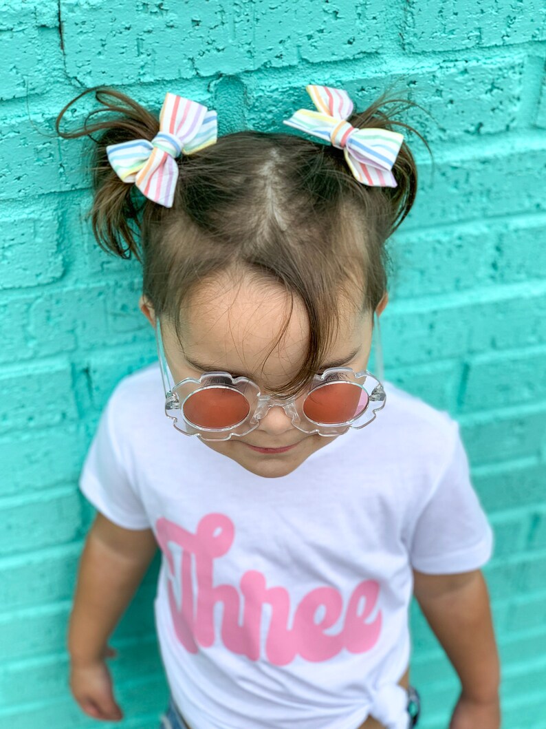 May include: A young child wearing a white t-shirt with the word "Three" in pink script. The child is wearing clear plastic sunglasses with red lenses and has rainbow striped bows in their hair.