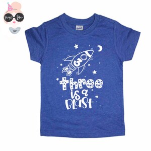 May include: A blue short-sleeved t-shirt with a white rocket ship and stars. The text "three is a blast" is printed on the shirt.