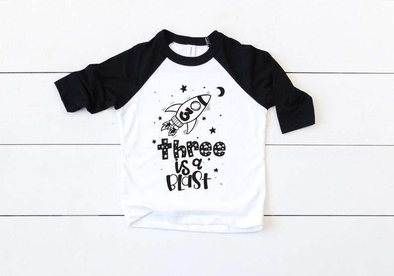 May include: A white and black baseball tee shirt with a black and white rocket ship graphic and the text "three is a blast".