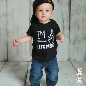 May include: A toddler wearing a black t-shirt with the text "I'm - Let's Party!" and a black mesh baseball cap. The toddler is wearing blue jeans and black and white sneakers.