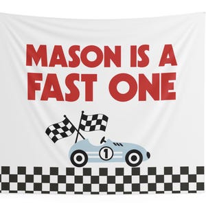 May include: White fabric wall hanging with the text "MASON IS A FAST ONE" in red. A light blue race car with the number 1 and checkered flags is featured below the text. A black and white checkered pattern borders the bottom.