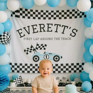May include: A baby sits in front of a backdrop with balloons and a banner that reads "EVERETT'S FIRST LAP AROUND THE TRACK." The banner features a race car graphic. The scene is decorated with checkered flags and a small blue cake.