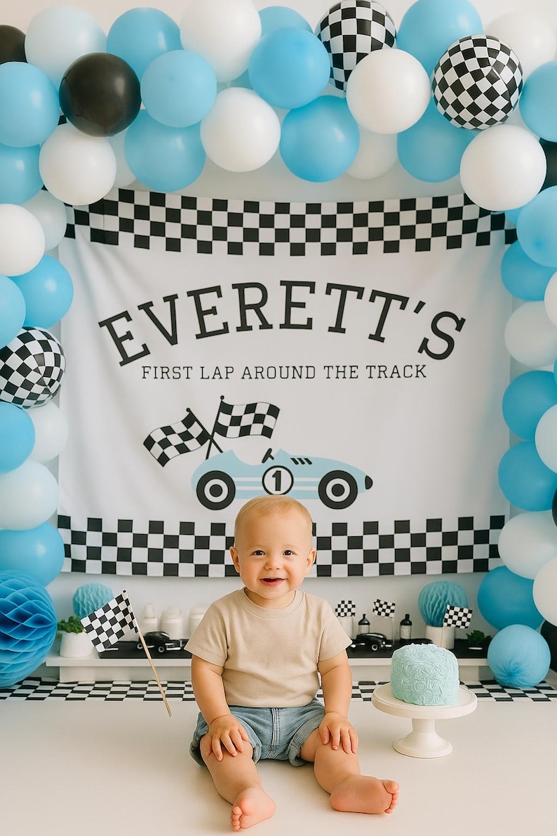 May include: A baby sits in front of a backdrop with balloons and a banner that reads "EVERETT'S FIRST LAP AROUND THE TRACK." The banner features a race car graphic. The scene is decorated with checkered flags and a small blue cake.