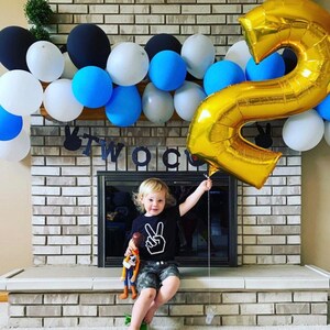 May include: A young child wearing a black shirt with a white peace sign graphic is sitting on a brick ledge. The child is holding a gold balloon shaped like the number two. The child is surrounded by a garland of balloons in various colours, including blue, black, and white. The garland is hanging above a brick fireplace. A black banner with the word "TWO" is hanging above the fireplace.