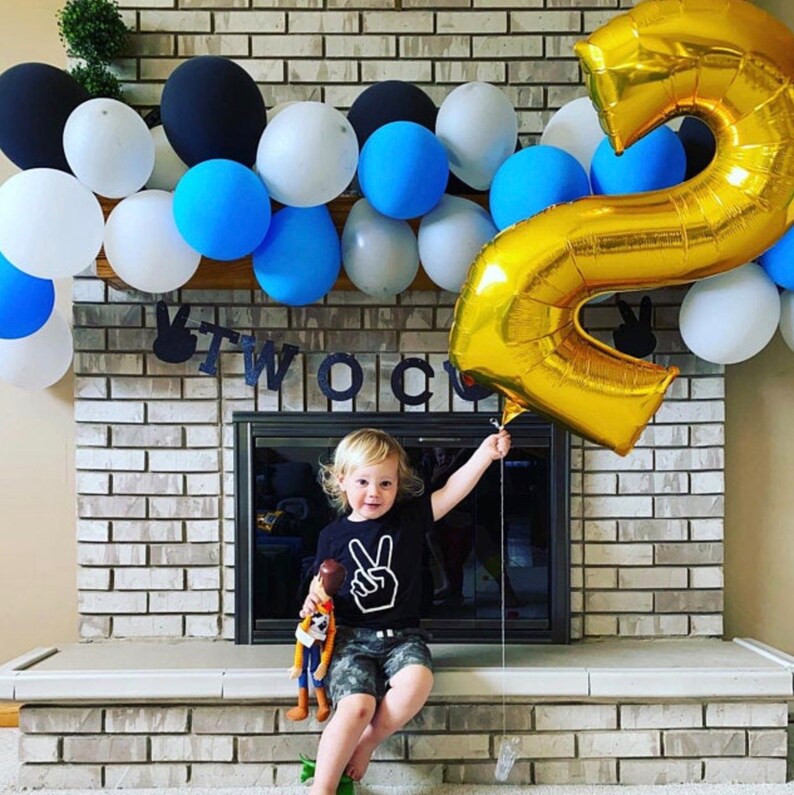 May include: A young child wearing a black shirt with a white peace sign graphic is sitting on a brick ledge. The child is holding a gold balloon shaped like the number two. The child is surrounded by a garland of balloons in various colours, including blue, black, and white. The garland is hanging above a brick fireplace. A black banner with the word "TWO" is hanging above the fireplace.