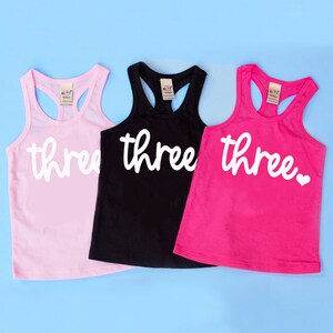 Puede incluir: Tres camisetas de tirantes en rosa, negro y rosa fuerte con la palabra "three" en letras blancas cursivas. La camiseta de tirantes rosa fuerte tiene un peque&ntilde;o coraz&oacute;n blanco despu&eacute;s de la palabra "three".