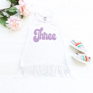 May include: White sleeveless dress with a purple "Three" graphic in a retro font. The dress has a fringe hem and is photographed on a white background.
