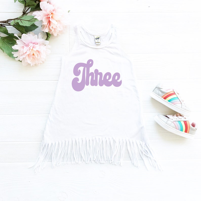 May include: White sleeveless dress with a purple "Three" graphic in a retro font. The dress has a fringe hem and is photographed on a white background.