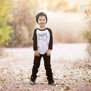 May include: A young boy wearing a black and white baseball tee shirt with the text "I'm" and a handprint graphic. He is wearing black pants and black shoes. The boy is standing in a path with fallen leaves on the ground.