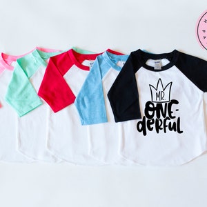 May include: Five baseball style shirts with different colored sleeves. The shirts are white with a black design that says "Mr. One-derful" with a crown above the text.