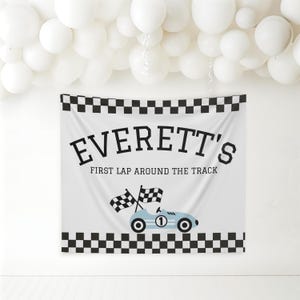 May include: White banner with black checkered borders and the text "EVERETT'S FIRST LAP AROUND THE TRACK". A light blue race car with the number 1 and checkered flags is featured below the text. White balloons are in the background.