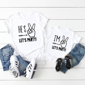 May include: Two white t-shirts with black text and graphics. The larger shirt says "He's Let's Party!" with a peace sign and arrow. The smaller shirt says "I'm Let's Party!" with a peace sign and arrow.