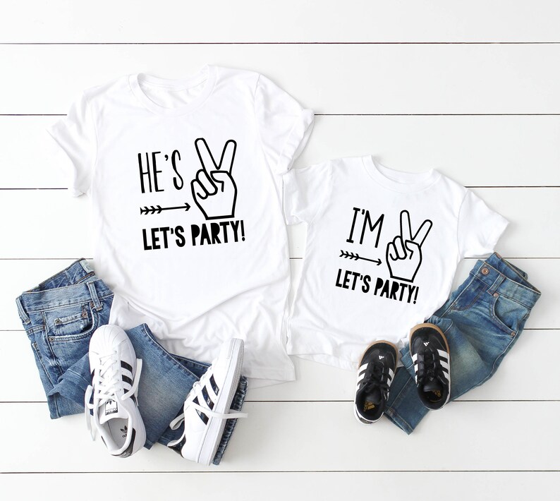 May include: Two white t-shirts with black text and graphics. The larger shirt says "He's Let's Party!" with a peace sign and arrow. The smaller shirt says "I'm Let's Party!" with a peace sign and arrow.