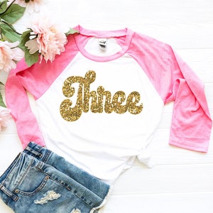 May include: A white and pink long-sleeved shirt with a gold glitter "Three" graphic. The shirt has a pink baseball style sleeve.