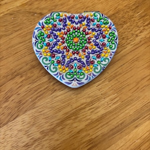 May include: A heart-shaped compact mirror with a colorful mandala design. The design features a variety of colors, including green, blue, purple, yellow, orange, and red. The mirror is surrounded by a white border.