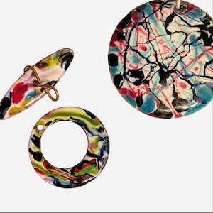 May include: Three decorative items featuring a marbled, multi-colored pattern of black, red, blue, green, and white. One item is a large circular pendant, another is a smaller circular ring, and the third is an elongated toggle with a wire loop.