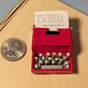 May include: A miniature red typewriter with white keys and a printed quote by Martin Luther King Jr. A U.S. quarter coin is next to the typewriter on a wooden surface. The quote reads: "Darkness cannot drive out darkness: only light can do that. Hate cannot drive out hate: Only love can do that."