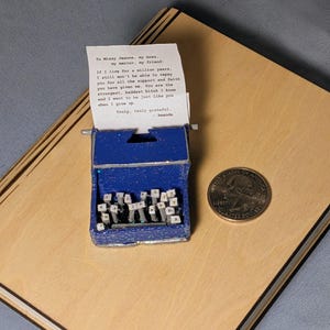 May include: A miniature blue typewriter with a handwritten note. The note reads, "To Missy Jeanne..." and includes a message of gratitude. A US quarter coin is next to the typewriter, which is on a wooden surface.