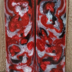 May include: Two rectangular resin blocks with a swirling pattern of red, black, and silver. The blocks have a glossy finish and are set against a wooden background. The design resembles a marbled effect.