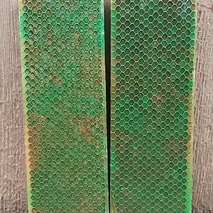 May include: Two rectangular bars of soap with a green honeycomb pattern overlaying a mix of green, yellow, and brown colors. The soap bars are displayed against a wooden background.