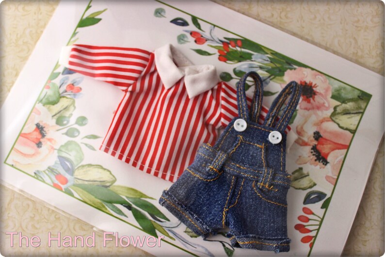 May include: Miniature doll clothing set featuring a red and white striped shirt with a white collar and denim overalls with white buttons. The clothing is displayed on a floral patterned background.