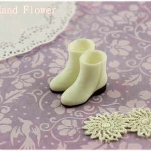 Puede incluir: Un par de botas blancas en miniatura con suelas marrones, junto a dos tapetes color crema, sobre una superficie lavanda con estampado floral. El texto "The Hand Flower" est&aacute; en la parte superior. Las botas son de color crema.