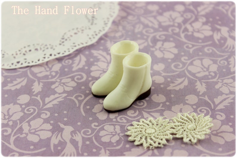 Puede incluir: Un par de botas blancas en miniatura con suelas marrones, junto a dos tapetes color crema, sobre una superficie lavanda con estampado floral. El texto "The Hand Flower" est&aacute; en la parte superior. Las botas son de color crema.