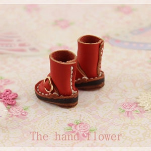 May include: A pair of miniature red leather boots with gold buckles and white stitching. The boots have brown soles and are set against a floral background. The text "The hand flower" is visible at the bottom.