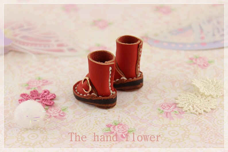 May include: A pair of miniature red leather boots with gold buckles and white stitching. The boots have brown soles and are set against a floral background. The text "The hand flower" is visible at the bottom.