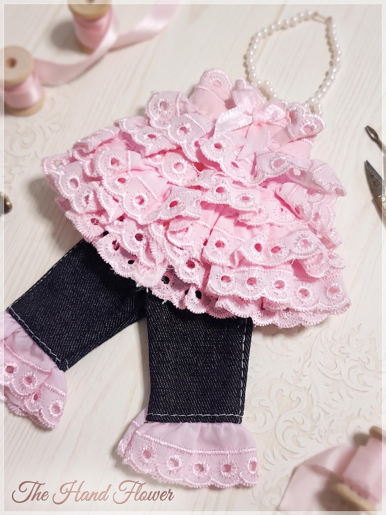 May include: A doll outfit consisting of a pink sleeveless top with multiple tiers of eyelet lace ruffles and a small bow, paired with dark blue denim trousers featuring matching pink eyelet lace trim at the cuffs. The text The Hand Flower is visible.