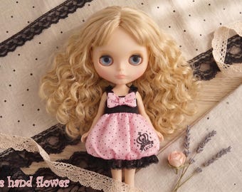 Op de afbeelding: Een Blythe-pop met blond krullend haar en blauwe ogen, gekleed in een roze polkadotjurk met een zwarte strik en afwerking. De pop staat op een gestippelde stof met kanten rand en de tekst "The hand flower".
