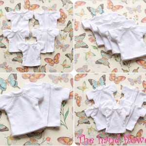 May include: Multiple white doll-sized t-shirts with short sleeves are arranged on a butterfly-patterned background. The shirts are made of a soft fabric and are neatly displayed, showcasing their simple design.