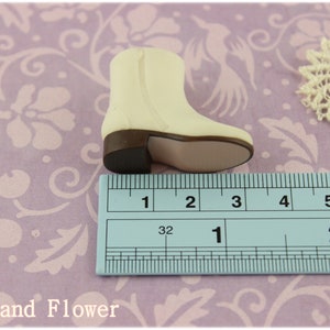 Puede incluir: Una bota en miniatura de color crema con tac&oacute;n y suela marrones, de aproximadamente 5 cm de largo, con cremallera. La bota est&aacute; sobre un fondo con estampado floral con un tapete de encaje.
