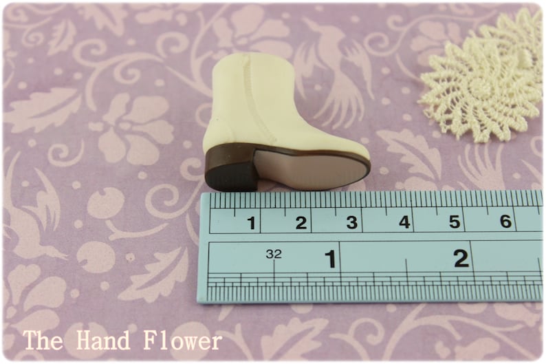 Puede incluir: Una bota en miniatura de color crema con tac&oacute;n y suela marrones, de aproximadamente 5 cm de largo, con cremallera. La bota est&aacute; sobre un fondo con estampado floral con un tapete de encaje.