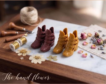 Puede incluir: Dos pares de botines en miniatura, un par en marrón oscuro y otro en amarillo mostaza, con múltiples correas con hebillas y tacones de bloque. Los botines se muestran junto a carretes de hilo dorado, herramientas y flores secas.