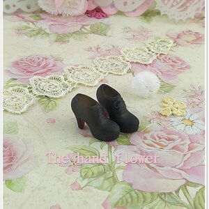 May include: A pair of miniature black lace-up shoes with high heels and red soles. The shoes are positioned on a surface featuring a floral pattern with pink roses and green leaves, accompanied by white lace trim and the text The hand flower.