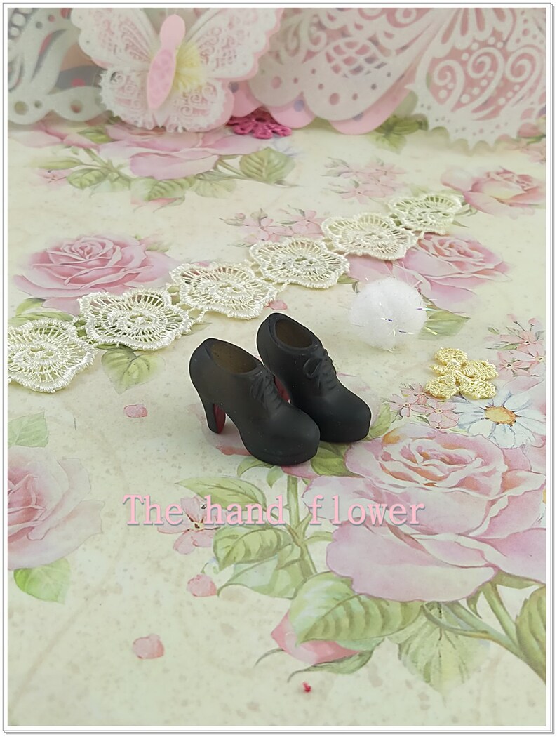 May include: A pair of miniature black lace-up shoes with high heels and red soles. The shoes are positioned on a surface featuring a floral pattern with pink roses and green leaves, accompanied by white lace trim and the text The hand flower.