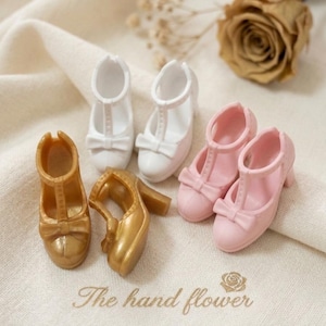 Op de afbeelding: Een verzameling miniatuur poppenschoenen in goud, wit en roze. De schoenen omvatten Mary Janes met strikken en sandalen met hakken. De tekst "The hand flower" is op de stof gedrukt.