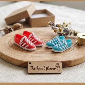 May include: Miniature red and blue sneakers with white stripes and laces are displayed on a wooden platter. The platter also holds a small spool of gold wire, dried flowers, and a small wooden tag that reads "The hand flower."