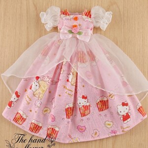 May include: A pink doll dress with a pattern of cupcakes and a cartoon cat. The dress has a white tulle overlay and puffy sleeves. A bow with a rose is at the waist. The text "The hand flower" is at the bottom.