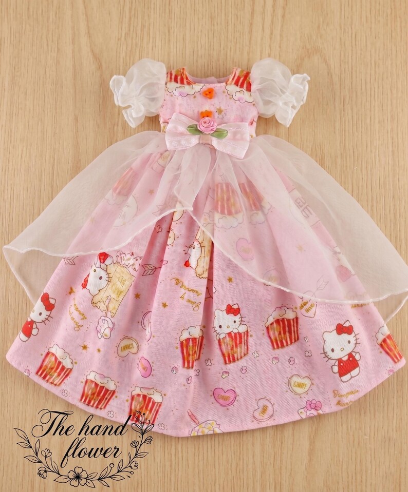 May include: A pink doll dress with a pattern of cupcakes and a cartoon cat. The dress has a white tulle overlay and puffy sleeves. A bow with a rose is at the waist. The text "The hand flower" is at the bottom.