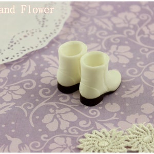Puede incluir: Dos peque&ntilde;as botas blancas con suelas marrones oscuras, sobre una superficie lavanda con estampado floral. Un tapete de encaje y el texto "The Hand Flower" tambi&eacute;n son visibles.