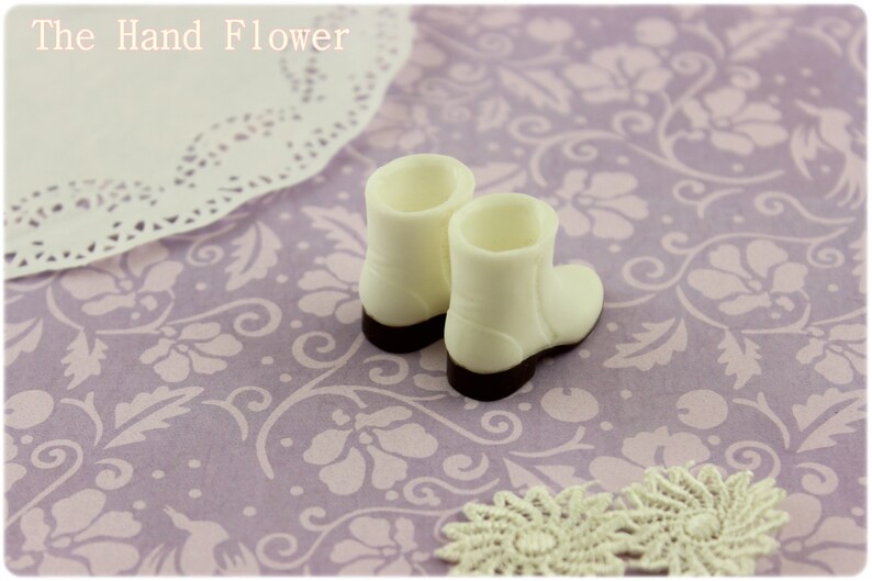 Puede incluir: Dos peque&ntilde;as botas blancas con suelas marrones oscuras, sobre una superficie lavanda con estampado floral. Un tapete de encaje y el texto "The Hand Flower" tambi&eacute;n son visibles.