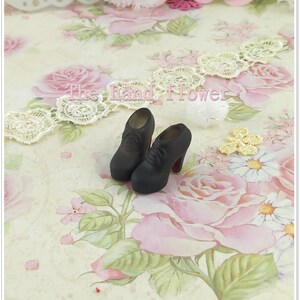 May include: A pair of miniature black lace-up high-heeled shoes with rounded toes and red soles. The shoes feature a smooth matte finish and are positioned on a surface with a floral pattern. The text "The hand flower" is visible in the center.